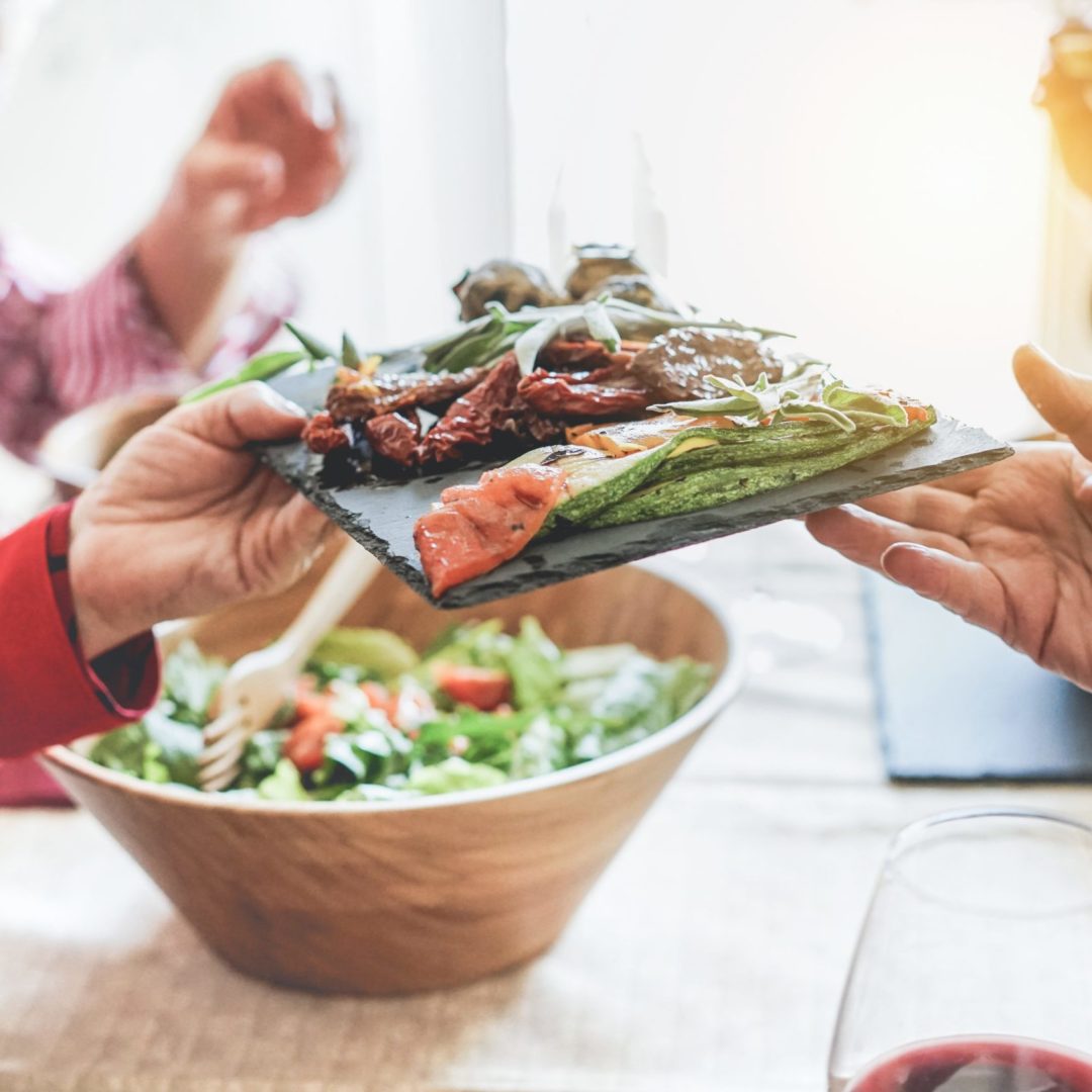 Senior people having a vegan lunch together. Photo shows hands passing a plate of vegetables from left to right. Plate includes roasted zucchini, tomatoes and salad. The person on the left is wearing a long-sleeved red jacket. In the background is a large salad bowl with salad. In the foreground is a glass of red wine.