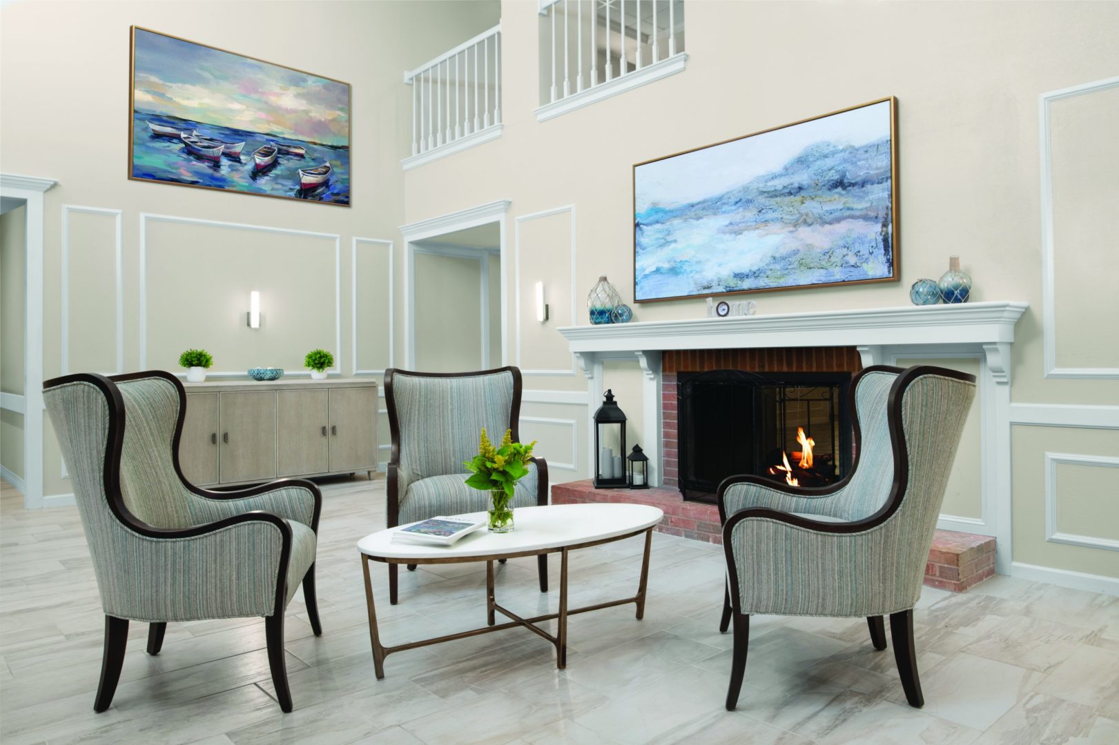 Front lobby and sitting area with three wing-backed chairs in front of a fireplace with warm fire. Three chairs surround a white, oval-shaped coffee table that holds two books and a vase with yellow flowers. The large fireplace mantle holds blue decorative glass jars and there is a large painting of blue mountains and water hanging above. On the far wall is a buffet-style storage cabinet with plants and a glass blue bowl. Above, on the wall, is a large painting of water with six unmanned row boats. The floor is beautiful large cream tiles. The space is open and bright.