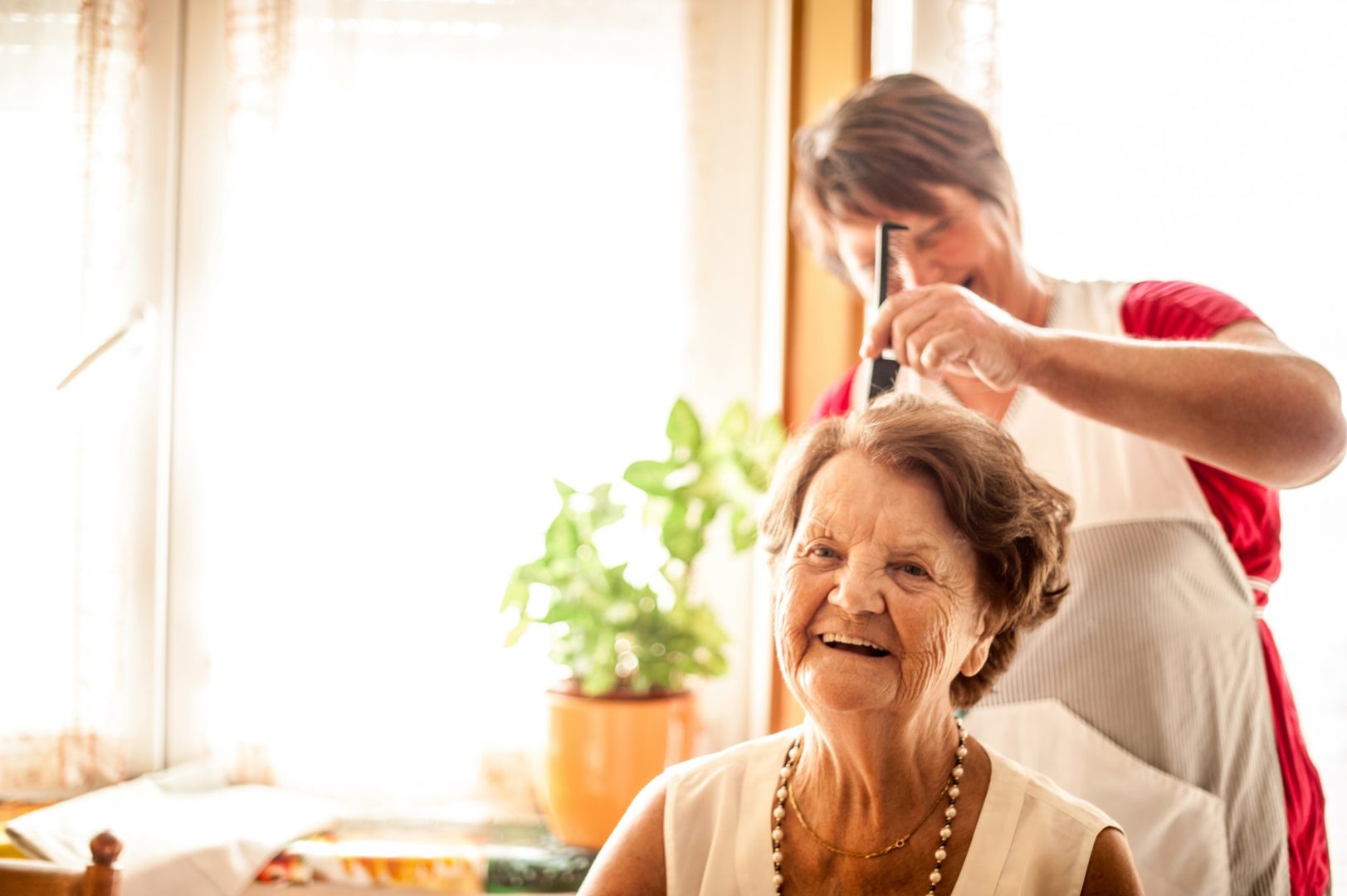 Caucasian senior woman has her hair styled. She is sitting in the foreground with the stylist to the back of her. She has short brown, wavy hair and is wearing a sleeveless, light cream linen top, a pearl necklace and short gold necklace. She is smiling. The stylist is behind her with a comb and is wearing a red top and apron. She has short brown hair. In the background is a large window and plant in a yellow pot.